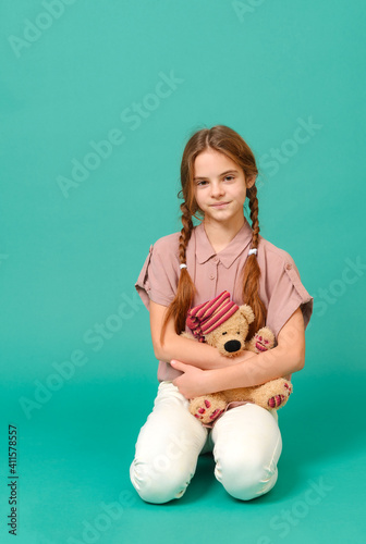 teenage girl in white trousers and a pink shirt sits with a toy bear on a green background