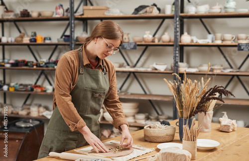 Portrait of young female artisan making plant imprint ceramics on wooden table in pottery workshop, small business and hobby concept, copy space