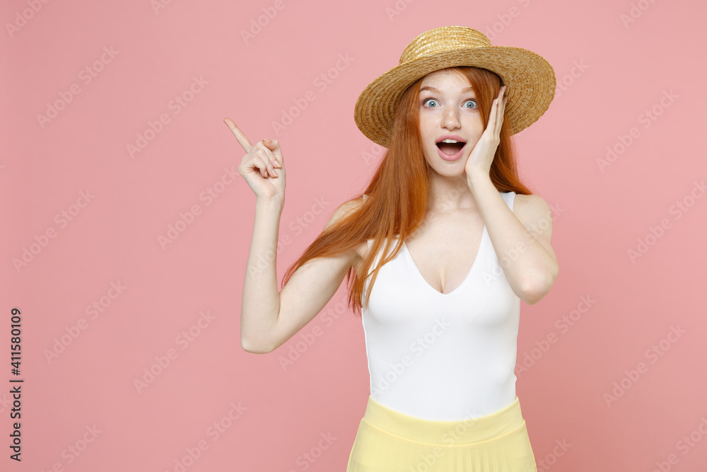 Young surprised shocked redhead woman ginger long hair in straw hat summer clothes point finger aside on copy space workspace mock up hold face isolated on pastel pink background studio portrait. Young surprised shocked redhead woman ginger long hair in straw hat summer clothes point finger aside on copy space workspace mock up hold face isolated on pastel pink background studio portrait.