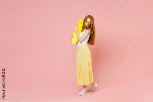 Full length body young smiling redhead energetic ginger teenager pretty caucasian woman 20s wear glasses summer clothes maxi skirt hold skateboard isolated on pastel pink background studio portrait.