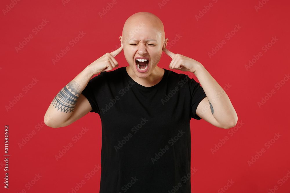 Fototapeta premium Young bald caucasian tired stressed tattooed woman 20s without hair wearing black basic casual t-shirt covering ear with fingers hands shout scream isolated on red color background studio portrait.
