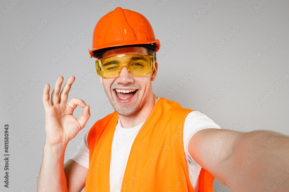 Young employee man in orange vest protective hardhat do selfie on mobile phone show ok okay gesture blink isolated on grey background studio Instruments for renovation apartment . Repair home concept. Young employee man in orange vest protective hardhat do selfie on mobile phone show ok okay gesture blink isolated on grey background studio Instruments for renovation apartment . Repair home concept.