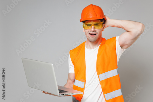 Young employee pensive man in orange vest protective helmet hold laptop pc computer sctratch back head isolated on grey background studio. Instruments for renovation apartment Repair home concept. photo