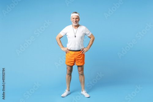 Full length elderly gray-haired sportsman fitness instructor smiling man 60s in sportswear white t-shirt whistle stand akimbo arm on waist isolated on blue background studio portrait. Sport concept.