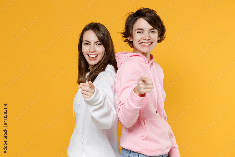 Cheerful smiling funny two young women friends 20s wearing casual white pink hoodies standing back to back pointing index fingers on camera isolated on bright yellow color background studio portrait. Cheerful smiling funny two young women friends 20s wearing casual white pink hoodies standing back to back pointing index fingers on camera isolated on bright yellow color background studio portrait.