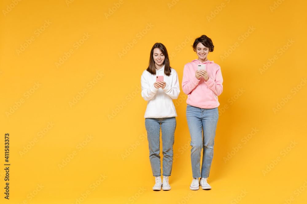 Full length of smiling attractive two young women friends 20s wearing casual white pink hoodies using mobile cell phone typing sms message isolated on bright yellow color background studio portrait. Full length of smiling attractive two young women friends 20s wearing casual white pink hoodies using mobile cell phone typing sms message isolated on bright yellow color background studio portrait.