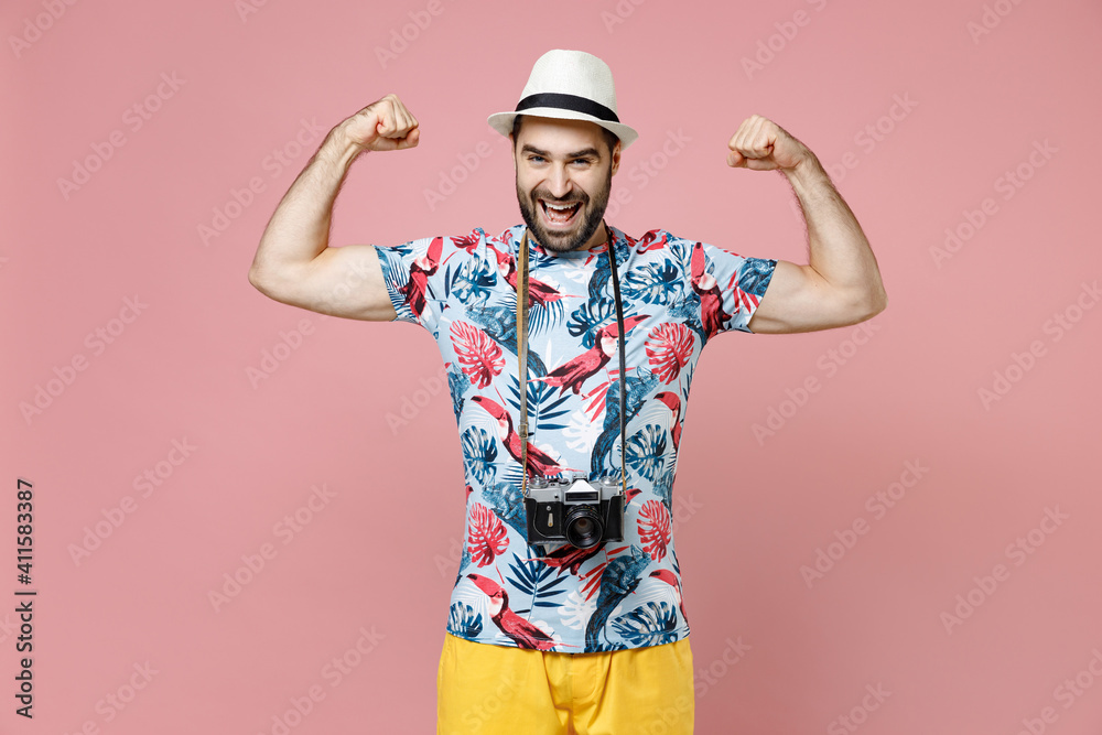 Strong young traveler tourist man in summer basic clothes hat with photo camera showing biceps muscles on hands isolated on pink background. Passenger traveling on weekend. Air flight journey concept.