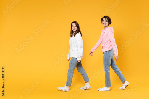 Full length side view of cheerful smiling funny two young women friends 20s wearing casual white pink hoodies walking going looking camera isolated on bright yellow color background studio portrait.