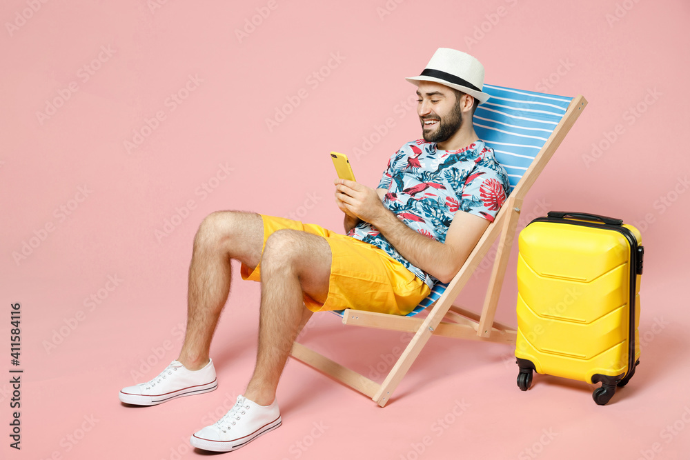 Full length smiling young traveler tourist man in hat sit on deck chair using mobile cell phone typing sms message isolated on pink background. Passenger travel on weekend. Air flight journey concept. - obrazy, fototapety, plakaty 