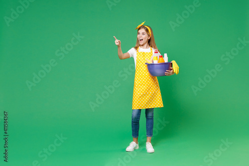 Full length excited little kid girl housewife 12-13 in apron point finger aside hold basin with detergent bottles washing cleansers doing housework isolated on green background. Housekeeping concept.