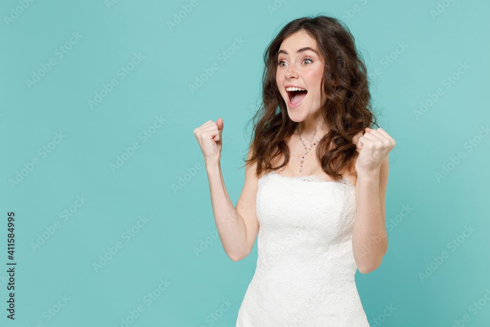 Excited bride young woman 20s in white wedding dress doing winner gesture celebrating clenching fist say yes isolated on blue turquoise background studio portrait. Ceremony celebration party concept.