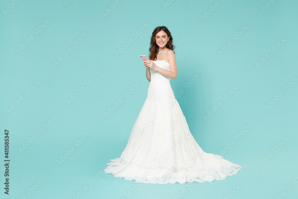 Full length of smiling bride young woman 20s in white wedding dress using mobile cell phone looking camera isolated on blue turquoise background studio portrait. Ceremony celebration party concept. Full length of smiling bride young woman 20s in white wedding dress using mobile cell phone looking camera isolated on blue turquoise background studio portrait. Ceremony celebration party concept.