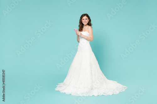 Full length of smiling bride young woman 20s in white wedding dress using mobile cell phone looking camera isolated on blue turquoise background studio portrait. Ceremony celebration party concept.