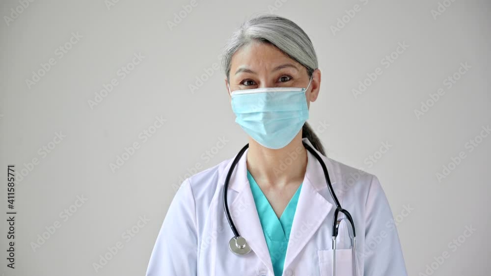 Close-up video portrait of asian gray-haired female doctor in medical uniform, protective mask and stethoscope stands against a white background and looks at the camera. Healthcare concept Close-up video portrait of asian gray-haired female doctor in medical uniform, protective mask and stethoscope stands against a white background and looks at the camera. Healthcare concept