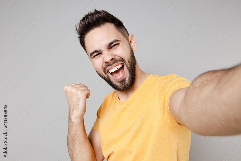 Close up young caucasian smiling bearded attractive man 20s in yellow basic t-shirt do selfie shot on mobile phone do winner gesture clench fist say yes isolated on grey background studio portrait. Close up young caucasian smiling bearded attractive man 20s in yellow basic t-shirt do selfie shot on mobile phone do winner gesture clench fist say yes isolated on grey background studio portrait.