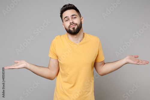 Young caucasian strict bearded handsome mistaken confused man 20s wearing casual yellow basic t-shirt spreading hands oops gesture looking camera isolated on grey color background studio portrait.