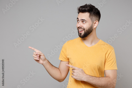 Young caucasian bearded smiling happy man in yellow basic t-shirt point index finger aside on workspace area copy space mock up isolated on grey background studio portrait People lifestyle concept