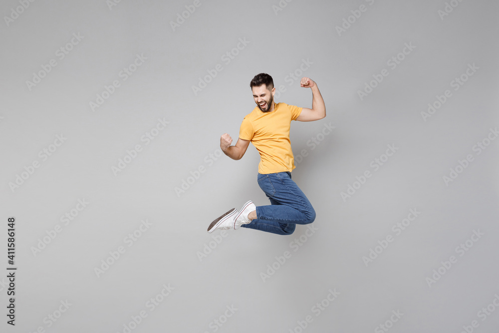 Full length of young bearded excited successful lucky overjoyed man 20s in yellow basic t-shirt walk do winner gesture clenching fist jump high celebrating isolated on grey background studio portrait