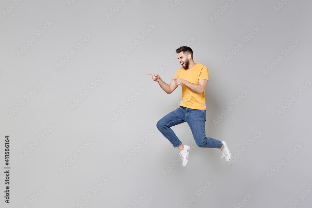 Full length of young bearded active student smiling overjoyed man 20s in yellow basic t-shirt jump high running away fast point index finger aside isolated on grey color background studio portrait..