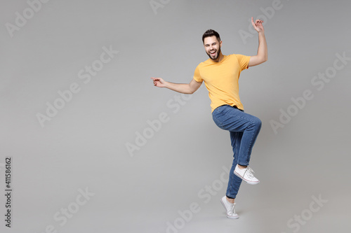 Full length of young bearded attractive smiling man 20s in casual yellow basic t-shirt with outstretched hand leaning over looking camera raised up leg isolated on grey background studio portrait.