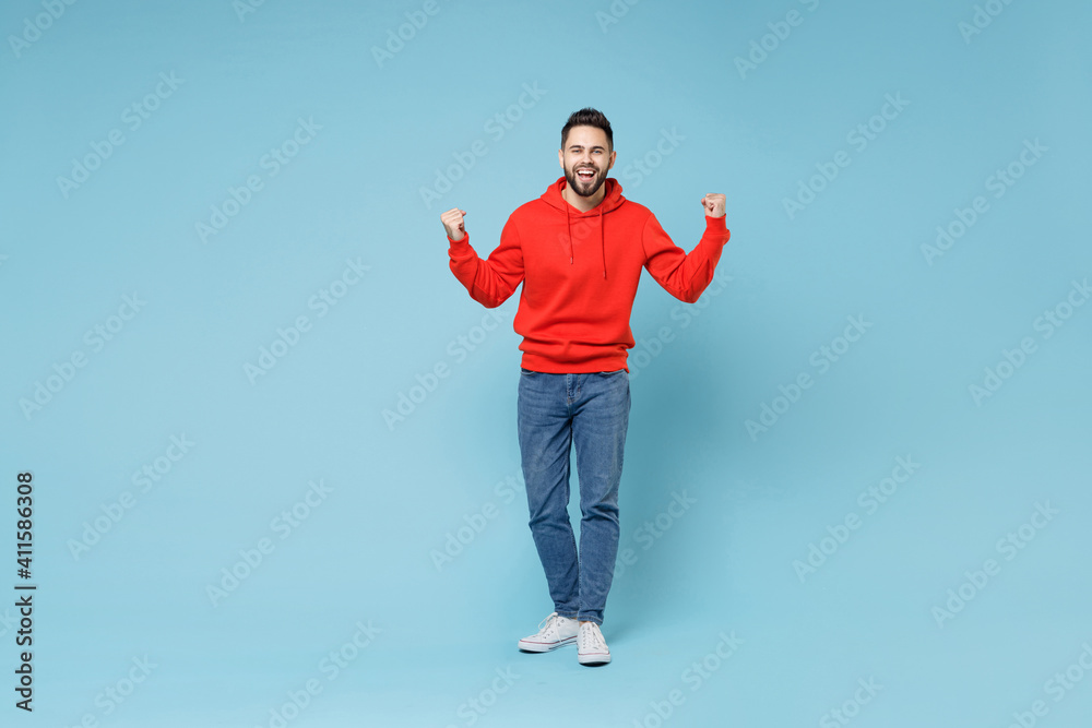 Full length of young caucasian happy bearded overjoyed excited man 20s in casual red orange hoodie doing winner gesture clenching fist celebrating isolated on blue color background studio portrait. Full length of young caucasian happy bearded overjoyed excited man 20s in casual red orange hoodie doing winner gesture clenching fist celebrating isolated on blue color background studio portrait.