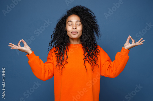 Smiling young african american woman wearing casual basic orange sweatshirt hold hands in yoga gesture, relaxing meditating, trying to calm down isolated on blue color wall background studio portrait.