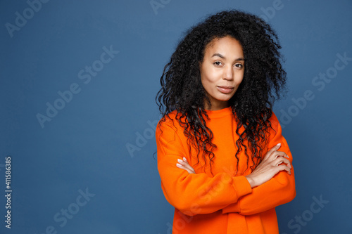 Smiling confident beautiful attractive young african american woman in casual basic bright orange sweatshirt holding hands crossed looking camera isolated on blue color background studio portrait. © ViDi Studio