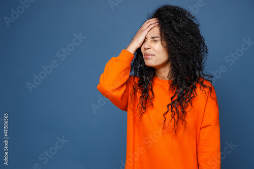 Schilderij op canvas Confused young african american woman in casual basic bright orange sweatshirt standing put hand on face facepalm epic fail gesture keep eyes closed isolated on blue color background studio portrait