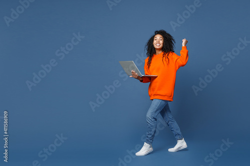 Full length side view of happy young african american woman wearing basic bright orange sweatshirt working on laptop pc computer doing winner gesture isolated on blue color background studio portrait.