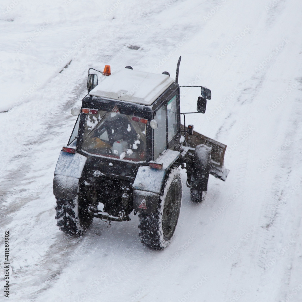 Old Belarus AWD wheeled tractor with a scraper shovel cleans snow on the street road after a snowfall on a winter day top rear view, city municipal machinery Old Belarus AWD wheeled tractor with a scraper shovel cleans snow on the street road after a snowfall on a winter day top rear view, city municipal machinery