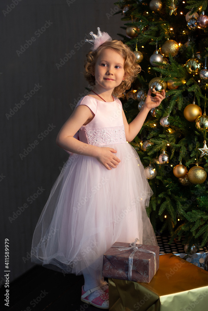 a little girl in a pink dress and a crown near the Christmas tree with gifts a little girl in a pink dress and a crown near the Christmas tree with gifts