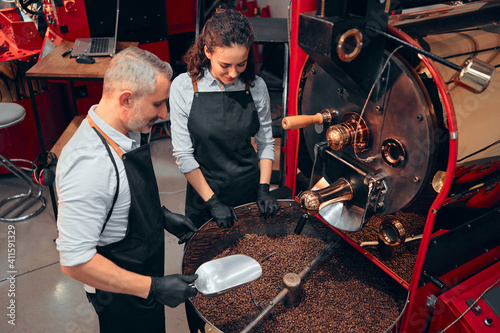 Slika na platnu Couple of two baristas checking the quality of the coffee beans standing with scoop near the roaster machine at the roastery