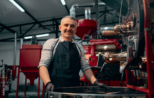 Portrait of smiling coffee roaster standing at roasting machine and looking at camera. Selective focus.