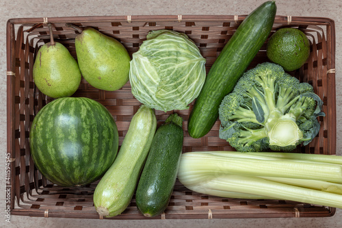 Flat lay composition of green fruits and vegetables including watermelon, pear, avocado, zucchini, cucumber, cabbage, broccoli, celery in brown rectangular basket