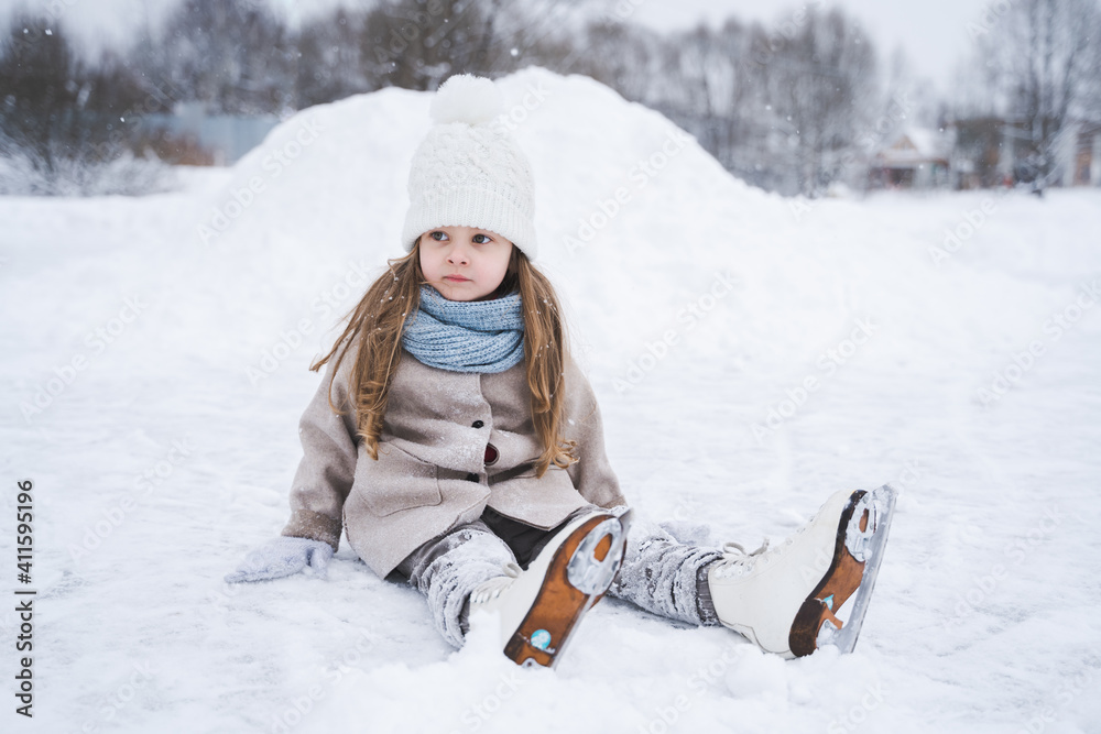 Fototapeta premium Beautiful little cute girl learn to skate on ice skating rink in park. Fall down and have fun. Stylish look, warm woolen coat, white hat, scarf, snood. Winter family activities, sport, games outdoors
