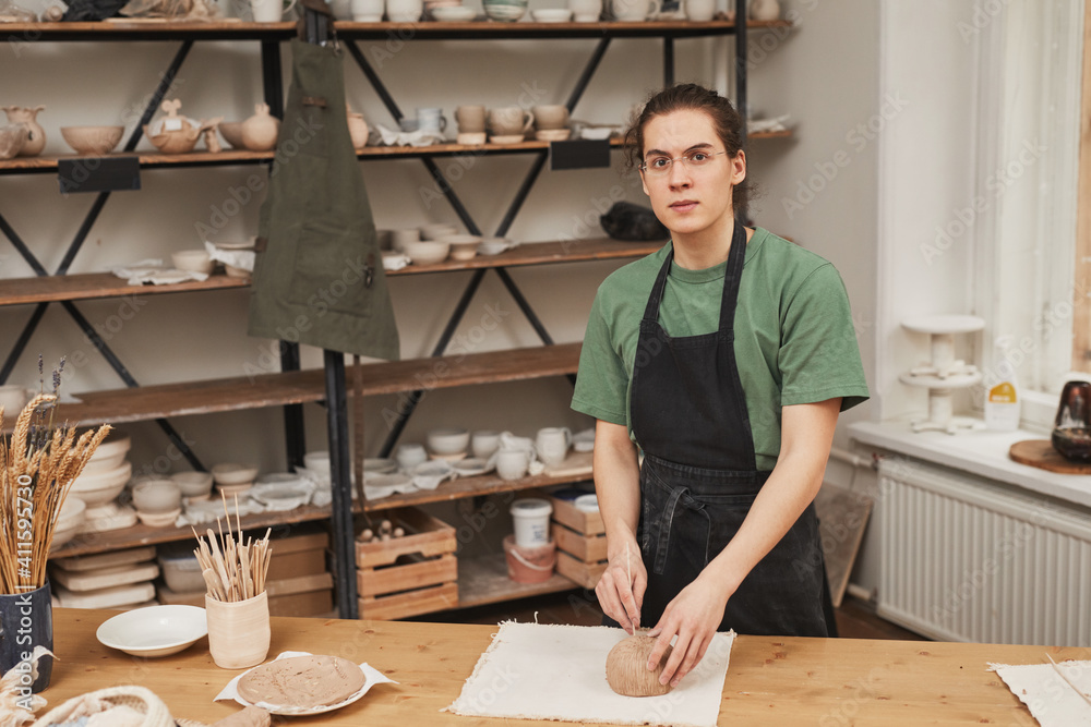Warm toned portrait of young man looking at camera while making ceramic bowl in pottery workshop, hobby and small business concept, copy space Warm toned portrait of young man looking at camera while making ceramic bowl in pottery workshop, hobby and small business concept, copy space