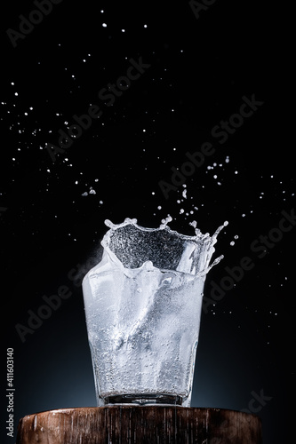 Pure mineral water in a glass, ice falls into the glass. Splashes of water rise above the glass, dark background with place for text, selective focus. Close-up, vertical frame