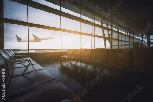 An empty dark modern waiting room of a contemporary airport terminal in the gate area, with rows of seats, during a lockdown, no people and a single white airplane gaining altitude outside the window