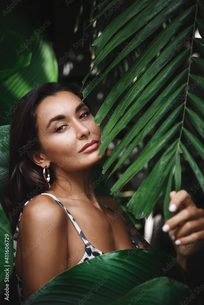 Close up of attractive young woman with perfect tan, wearing bikini, posing in tropical jungle palm leaves with sensual smile. Female tourist relaxing on vacation Close up of attractive young woman with perfect tan, wearing bikini, posing in tropical jungle palm leaves with sensual smile. Female tourist relaxing on vacation