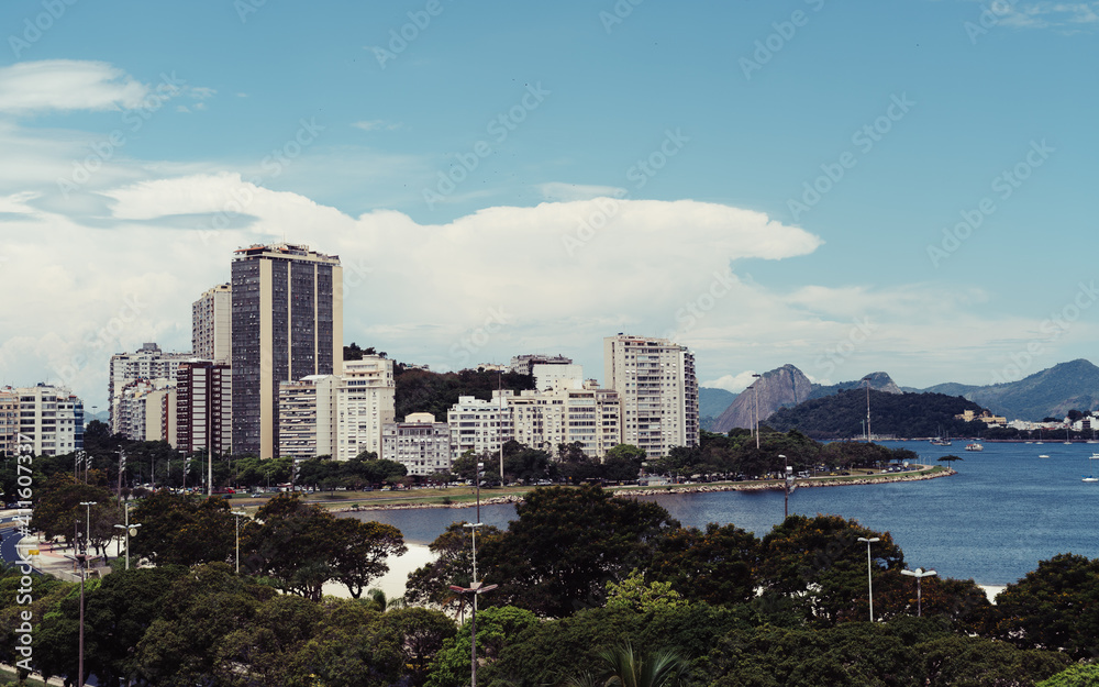 An urban cityscape with a group of trees surrounding the road in the foreground and multiple residential buildings near the bay with several sailboats and far hills and mountains in the background An urban cityscape with a group of trees surrounding the road in the foreground and multiple residential buildings near the bay with several sailboats and far hills and mountains in the background