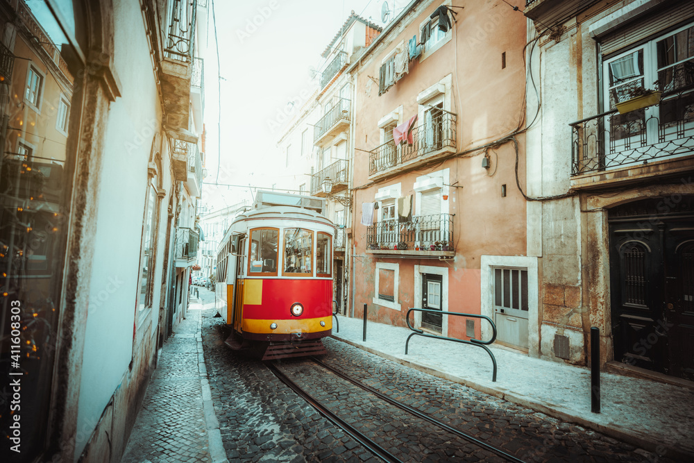 A wide-angle view of a red retro tram on a narrow street with one-way rail traffic in a European city; a vintage tourist streetcar in yellow and red colors on a tramway track over paving stone