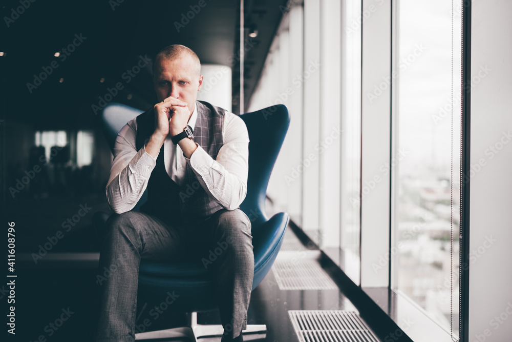 A portrait of concentrated hard thinking nervous man entrepreneur with hands closing his mouth sitting on a blue armchair next to the window of a luxury business high-rise, considering a crisis plan A portrait of concentrated hard thinking nervous man entrepreneur with hands closing his mouth sitting on a blue armchair next to the window of a luxury business high-rise, considering a crisis plan