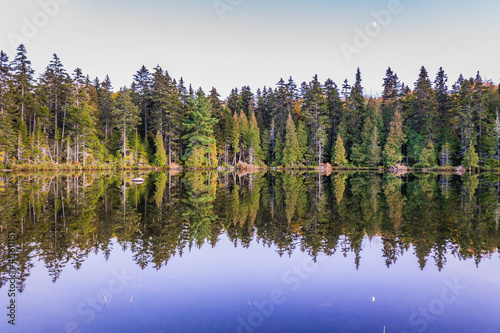 The sun is setting and the pine trees are reflecting in the lake in Mauricie National Park, a national park located in Quebec, Canada © Pernelle Voyage