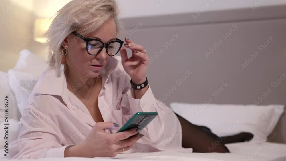 A blonde woman with glasses is in the bedroom, lying on her stomach on a bed with white linens and looking at her smartphone. A blonde woman with glasses is in the bedroom, lying on her stomach on a bed with white linens and looking at her smartphone.