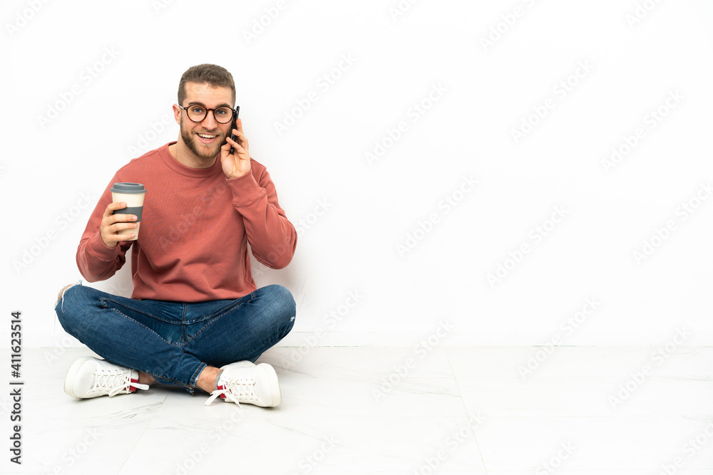 Young handsome man sitting on the floor holding coffee to take away and a mobile Young handsome man sitting on the floor holding coffee to take away and a mobile