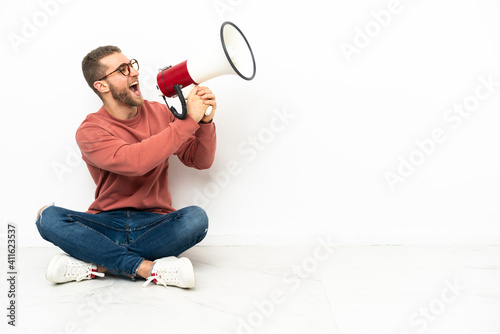 Young handsome man sitting on the floor shouting through a megaphone