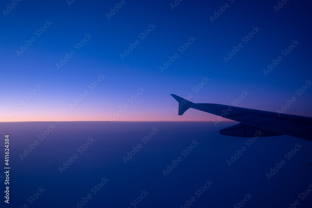 The left side wing of a large white commercial airplane flying over the the Atlantic Ocean under the morning sun. The horizon is pink from the sunrise, the sky is blue and the ocean is dark blue. The left side wing of a large white commercial airplane flying over the the Atlantic Ocean under the morning sun. The horizon is pink from the sunrise, the sky is blue and the ocean is dark blue.