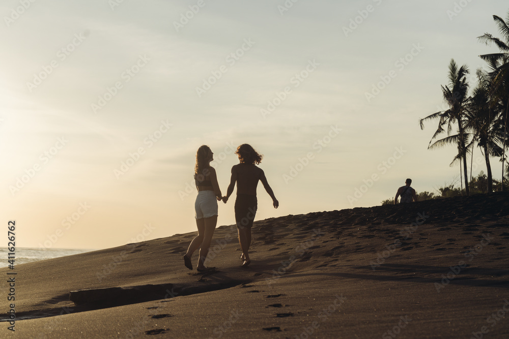 A young couple of lovers runs on a tropical beach against the backdrop of a sunset and holds hands. High quality photo