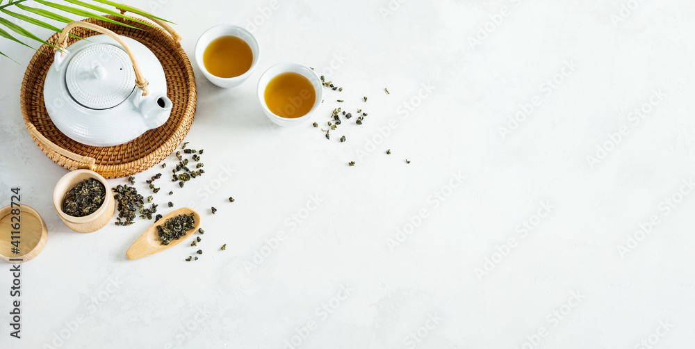 Asian tea concept, two white cups of tea and teapot surrounded with green dry tea  with space for a text on light background. Wide banner. - obrazy, fototapety, plakaty 