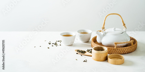 Asian tea concept mockup, two white cups of tea and teapot surrounded with green dry tea  with space for a text on light background. Wide banner.
